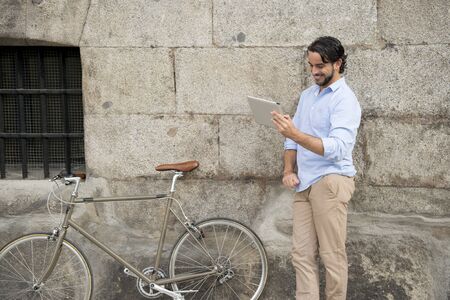 young happy attractive hispanic man in casual trendy clothes smiling cheerful using internet with digital tablet pad on vintage cool retro bicycle in modern stylish lifestyle urban style concept の写真素材