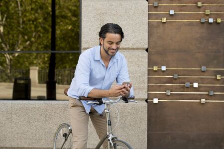 young happy attractive hispanic man in casual trendy clothes smiling cheerful using mobile phone on vintage cool retro bicycle in modern stylish lifestyle urban style concept の写真素材