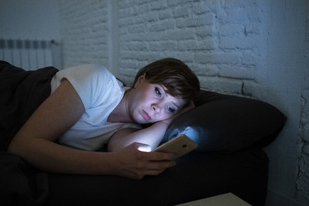 Young attractive woman awake late at night using smart phone lying in bed in a dark bedroom. Using...
