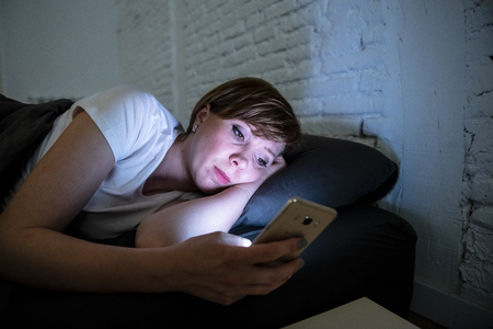Young attractive woman awake late at night using smart phone lying in bed in a dark bedroom. Using mobile for chatting and sending messages in internet addiction, mobile abuse and insomnia conceptの写真素材