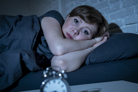Young attractive red haired caucasian woman lying in bed late at night trying to sleep suffering from insomnia, nightmares or sleeping disorders. looking stressed and exhausted.の写真素材