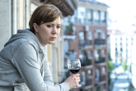 Beautiful red haired caucasian woman suffering from depression holding a glass of wine on a balcony...