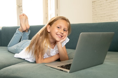 Beautiful cute charming little blonde girl playing and surfing the internet on laptop smiling and lying down on the couch at home. Digital technology, internet usage and life style conceptの写真素材