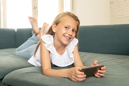 Beautiful cute charming little blonde girl playing and having fun on her smart phone smiling and...