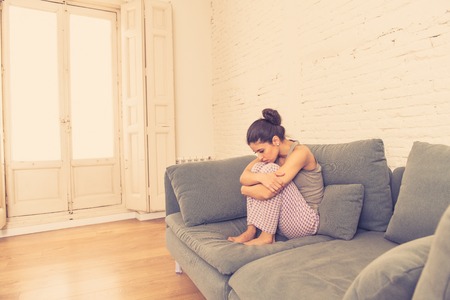 Beautiful latin depressed lonely woman staring out feeling sad, pain, grief at home on her sofa. Crisis, depression and mental health conceptの写真素材