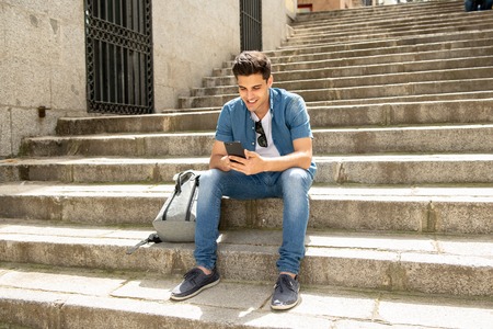 young happy student man in jeans denim clothes wearing sunglasses texting on his smart mobile phone and smiling, outside on the street of a modern european city in communication conceptの写真素材