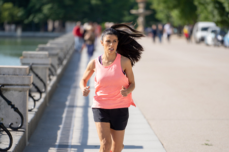 Running woman runner suffering from jogging in sunny bright light female fitness model training outside in city green park crossing training in fitness exercise conceptの写真素材
