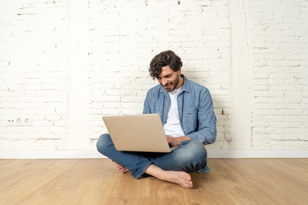portrait of latin man dressed in T-shirt blue shirt and jeans sitting against white brick wall holding laptop and watching media with happy smile in hipster freelance working from home concept.の写真素材