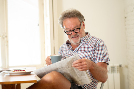 Portrait of happy senior old man with grey hair reading the newspaper while having breakfast and drinking a coffee in happy old age retirement concept.のeditorial素材