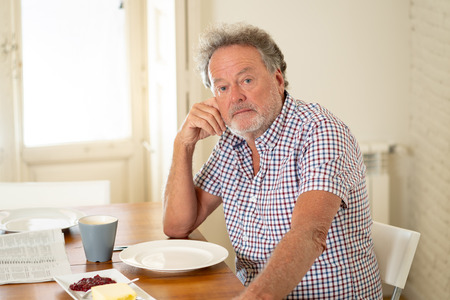 Portrait of depressed senior old man with grey hair reading the newspaper while having breakfast and drinking a coffee in old age retirement concept.の写真素材