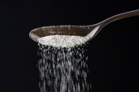 Studio picture of a metal spoon pouring sugar isolated on a black background in cooking sweet sugar addiction unhealthy diet overweight and obesity conceptの写真素材