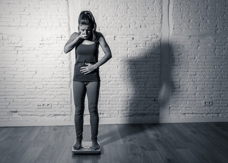 Young fit and slim woman checking body weight on scale finding herself fat and feeling depressed and desperate with big edgy shadow light in eating disorder and dieting concept.の写真素材
