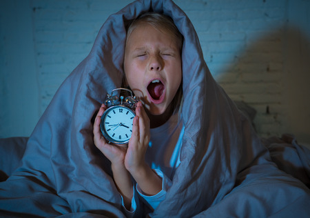 Cute sleepless little girl lying in bed showing alarm clock looking tired having sleeping troubles staying asleep at night or waking too early in Children Insomnia Anxiety and Sleep Disorders.の写真素材