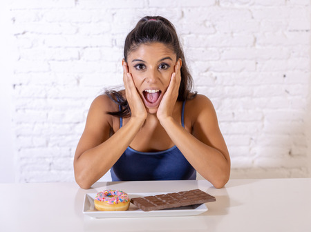 Young attractive latin woman sitting at table about to eat chocolate and doughnuts looking excited and happy in no more diet, sugar and chunky unhealthy food concept.の写真素材