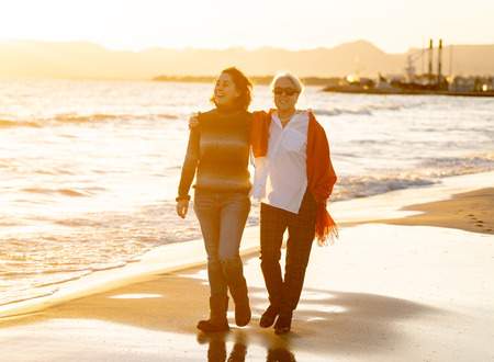 Portrait of happy senior mother and adult daughter spending time together holding hands laughing and walking on beach at sunset light in Happy family moments Generations Retirement and People concept.の写真素材