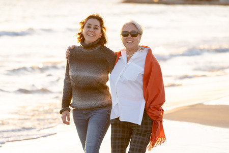 Portrait of happy senior mother and adult daughter spending time together holding hands laughing and walking on beach at sunset light in Happy family moments Generations Retirement and People concept.の写真素材