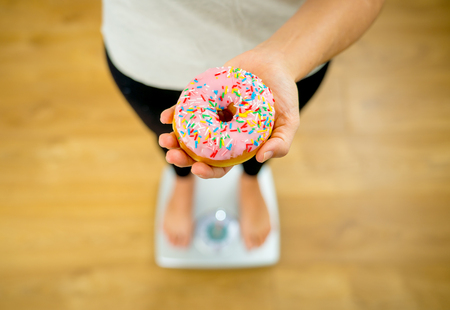 Close up of woman on scale holding doughnut on hand unhappy of body weight and eating habits of too much junk unhealthy food on Diet in Nutrition Detox Weight loss and Healthy lifestyle concept.の写真素材