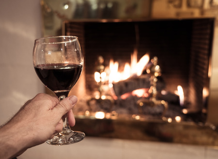 Close up of man holding and drinking red wine glass by open wood fire place relaxing and enjoying quiet moment at home in winter holiday vacation, Christmas or leisure concept.の写真素材