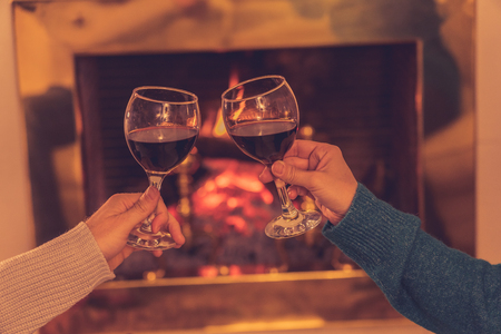 Close up of romantic couple holding two red wine glasses toasting at fire celebrating happy moment together by fireplace at cabin home in winter holiday vacation, Christmas or anniversary concept.の写真素材