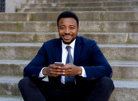 Happy african american businessman on mobile phone checking text messages social network sitting outside city stairs in People, technology online connection and entrepreneur creative business concept.の写真素材