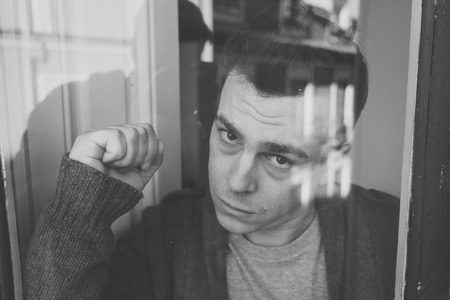 Close up portrait of young sad unhappy caucasian man looking worried, desperate and suicidal through the window at home. feeling worthless and in pain. In People, Depression and crisis concept.の写真素材
