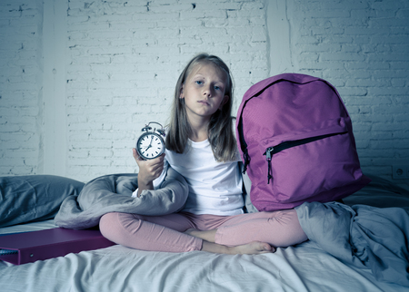 Beautiful blonde little girl sad sleepless and angry showing alarm clock time to get ready for school in difficulties waking up in the morning Children insomnia and sleeping disorders concept.の写真素材