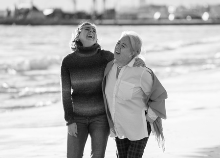 Portrait of happy senior mother and adult daughter spending time together holding hands laughing and walking on beach at sunset light in Happy family moments Generations Retirement and People concept.の写真素材