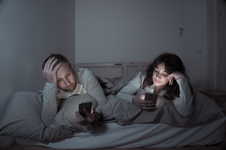 Life style portrait of young bored couple in bed at night on smart phones obsessed with games, social media, apps ignoring each other. Relationship communication problems and phone addiction concept.の写真素材