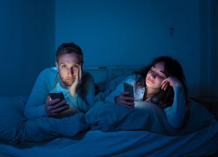 Life style portrait of young bored couple in bed at night on smart phones obsessed with games,...