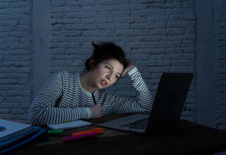 Overworked and tired female student working late at night on her laptop trying not to fall asleep feeling fatigued, worried and sad. Moody dark light. Online learning and stress work concept.の写真素材