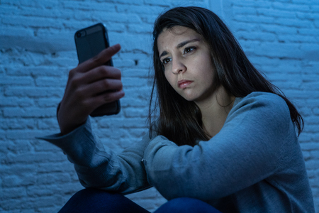 Portrait of sad scared young woman on the floor at night stressed and worried on smart mobile phone suffering cyber bullying harassment. desperate and depressed by being online abused by stalker.の写真素材