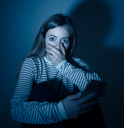 Sad desperate young teenager female girl on smart phone suffering from online bulling and harassment felling lonely and hopeless sitting on bed at night. CYberbullying and dangers of internet concept.の写真素材
