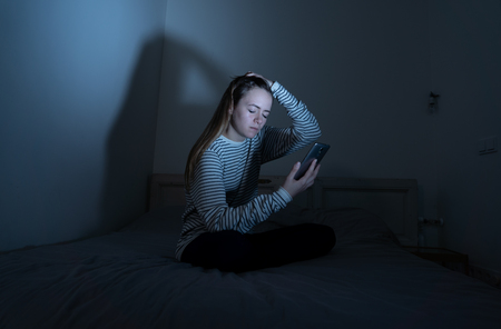 Sad desperate young teenager female girl on smart phone suffering from online bulling and harassment felling lonely and hopeless sitting on bed at night. CYberbullying and dangers of internet concept.の写真素材