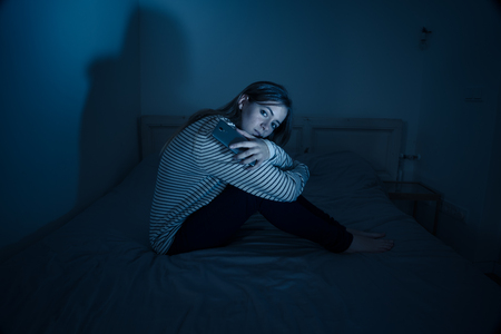 Young teenager girl suffering mobile cell phone addiction feeling lonely and depressed having insomnia needing to be connected sitting on bed late at night. Inの写真素材
