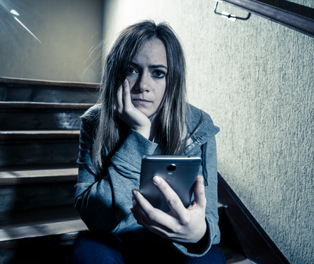 Sad depressed young teenager girl victim of cyberbullying by mobile smart phone siting on stairs feeling lonely, unhappy, hopeless and abused. Bullied by text message on social media app. Dark lightの写真素材