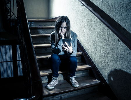 Teenager girl victim of online stalker suffering from cyberbullying abuse feeling lonely and hopeless sitting on stairs with dark light. Dangers of internet, online grooming and harassment concept.の写真素材