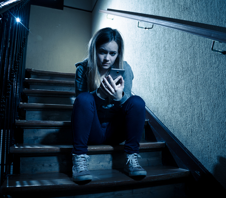 Sad depressed young teenager girl victim of cyberbullying by mobile smart phone siting on stairs feeling lonely, unhappy, hopeless and abused. Bullied by text message on social media app. Dark lightの写真素材