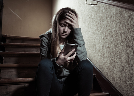 Teenager girl victim of online stalker suffering from cyberbullying abuse feeling lonely and hopeless sitting on stairs with dark light. Dangers of internet, online grooming and harassment concept.の写真素材