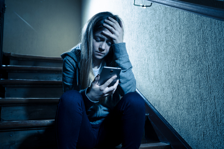 Teenager girl victim of online stalker suffering from cyberbullying abuse feeling lonely and hopeless sitting on stairs with dark light. Dangers of internet, online grooming and harassment concept.の写真素材