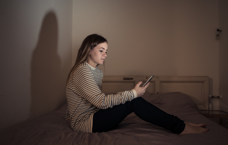 Young teenager girl suffering mobile cell phone addiction feeling lonely and depressed having insomnia needing to be connected sitting on bed late at night. Inの写真素材