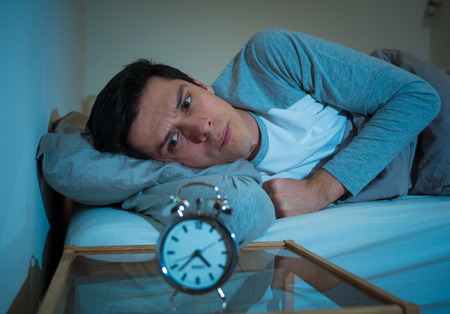 Sleepless and desperate young caucasian man awake at night not able to sleep, feeling frustrated and worried looking at clock suffering from insomnia in stress and sleep disorder concept.の写真素材
