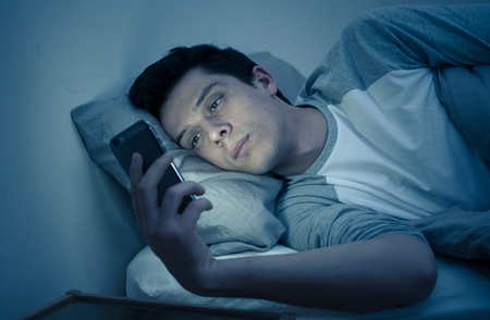 Addicted man chatting and surfing on the Internet with smart phone late at night in bed. Bored, sleepless and tired in dark room with moody light. In insomnia and mobile addiction concept.の写真素材