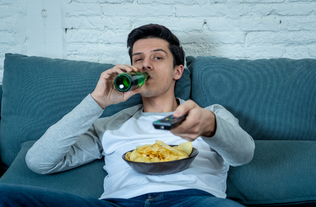 Lifestyle portrait of young bored man on couch with remote control zapping for movie or live sport. Looking disinterested drinking beer. Sedentary and mass social media or Television addiction.の写真素材