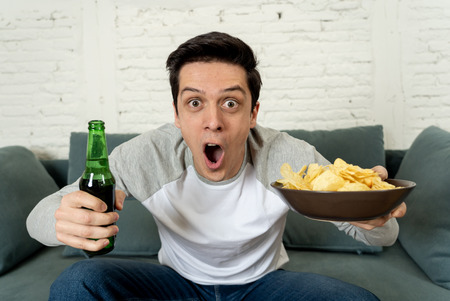 Lifestyle portrait of young man on sofa watching sports or exciting movie on television. Having fun at home enjoying and celebrating goal and victory drinking beer. In entertainment and mass media.の写真素材