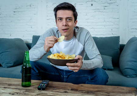 Lifestyle portrait of young man on sofa watching sports or exciting movie on television. Having fun at home enjoying and celebrating goal and victory drinking beer. In entertainment and mass media.の写真素材
