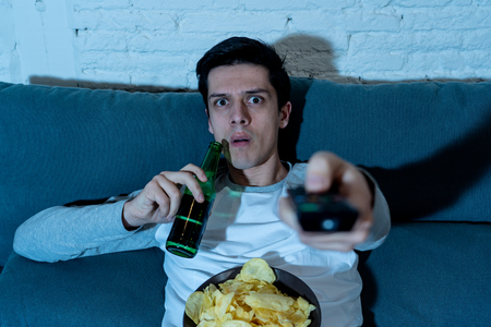 Lifestyle portrait of young man feeling scared and shocked making fear, anxiety gestures while watching TV, holding remote control and drinking a beer and eating snacks. In mass media and leisure.の写真素材