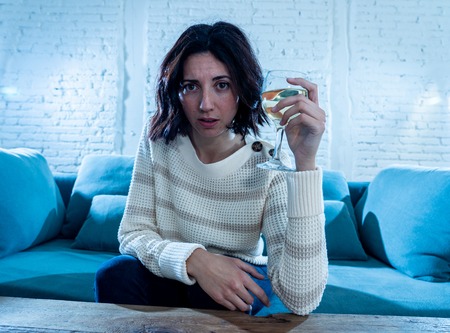 Portrait of depressed woman drinking glass of wine alone at home. Feeling distress, hopeless and frustrated, trying to feel better drinking. Unhealthy behavior, depression and alcohol concept.の写真素材