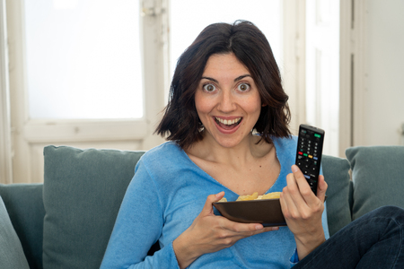 Happy woman on sofa with TV remote control ready to watch favorite movie of TV Show. Looking enthusiastic, making gestures of approval and eating chips. In people, technology and leisure concept.の写真素材