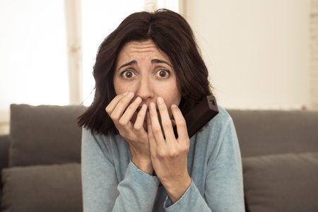 Lifestyle portrait of woman feeling scared and shocked making fear, anxiety gestures while watching horror movie on TV holding remote control. In horror and violence on TV and internet and mass media.の写真素材