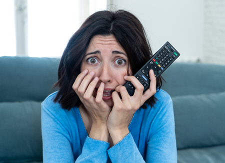 Lifestyle portrait of woman feeling scared and shocked making fear, anxiety gestures while watching horror movie on TV holding remote control. In horror and violence on TV and internet and mass media.の写真素材